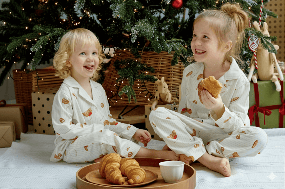 Two kids in matching Christmas pajamas enjoying croissants under the Christmas tree