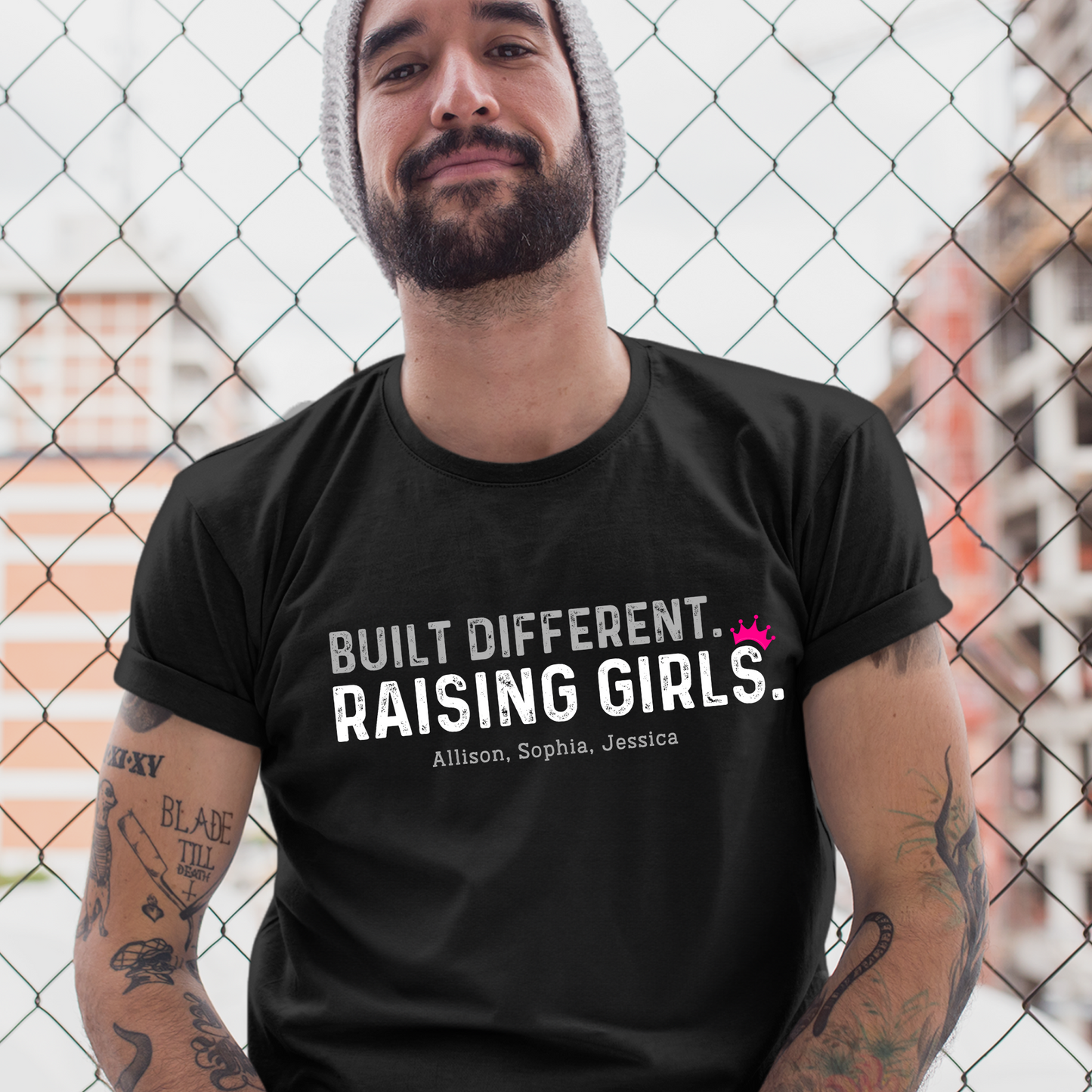 Man wearing a black t-shirt that reads “Built Different. Raising Girls.” with custom daughter names below. Shirt is styled with rolled sleeves and photographed outdoors in front of a wire fence.