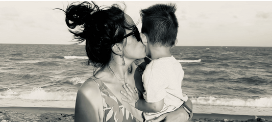 Mother holding toddler and kissing his cheek while standing at the beach, soft and intimate moment.
