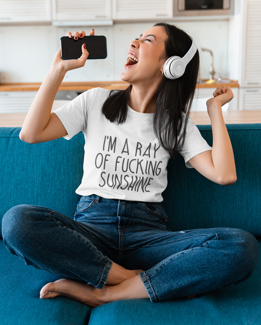 Mockup of a young woman sitting on a blue couch, wearing a white t-shirt with bold text that reads 'I'm a ray of f***ing sunshine,' laughing joyfully while holding a smartphone and wearing headphones.