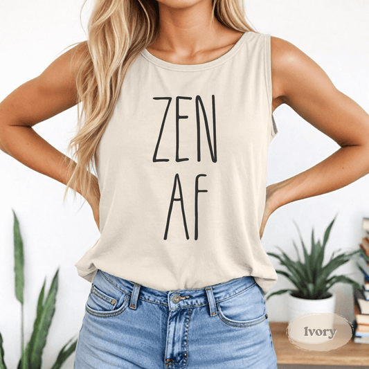 Woman wearing an ivory tank top with minimalist black text that reads “ZEN AF,” standing casually with one hand on her hip and plants in the background.