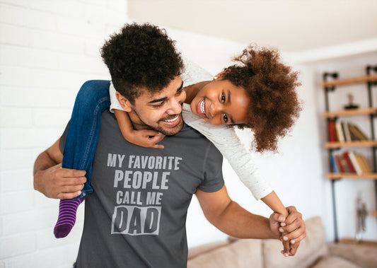 Smiling dad carrying his young daughter on his back while wearing a charcoal gray T-shirt that says “My Favorite People Call Me Dad” in bold white lettering.

