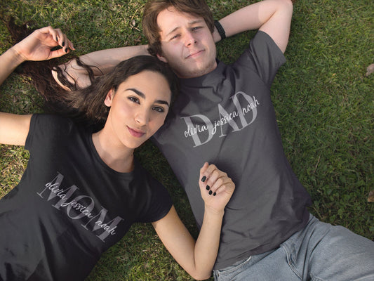 Couple lying on grass wearing matching personalized MOM and DAD shirts with kids' names printed across the chest in elegant script font.

