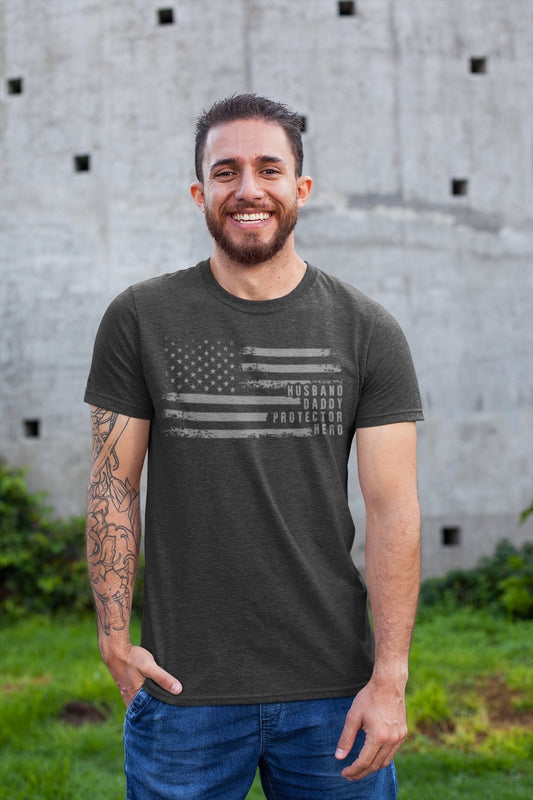Man smiling and wearing a charcoal t-shirt that features a distressed American flag and the words "Husband. Daddy. Protector. Hero." — a patriotic Father's Day gift shirt.

