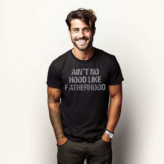 Smiling man wearing a black “Ain’t No Hood Like Fatherhood” T-shirt, standing confidently with hands in pockets.

