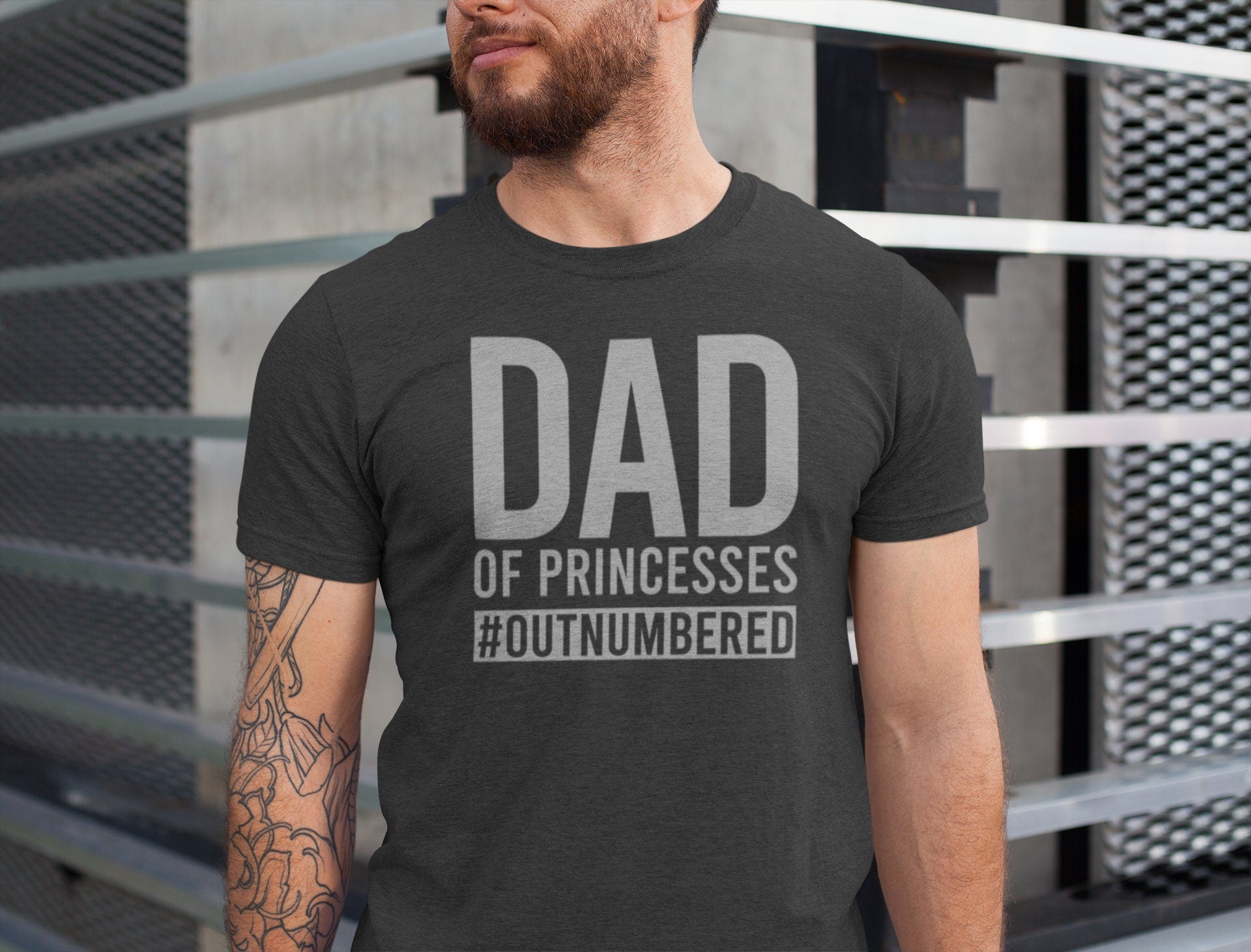 Man wearing a dark gray “Dad of Princesses #Outnumbered” T-shirt, standing by a modern metal structure.


