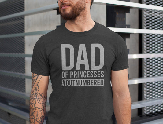 Man wearing a dark gray “Dad of Princesses #Outnumbered” T-shirt, standing by a modern metal structure.

