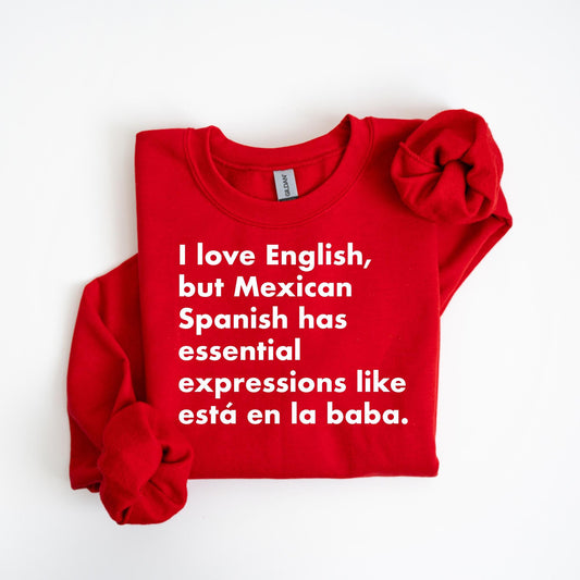 Red folded Gildan sweatshirt with white text that reads: “I love English, but Mexican Spanish has essential expressions like está en la baba.”

