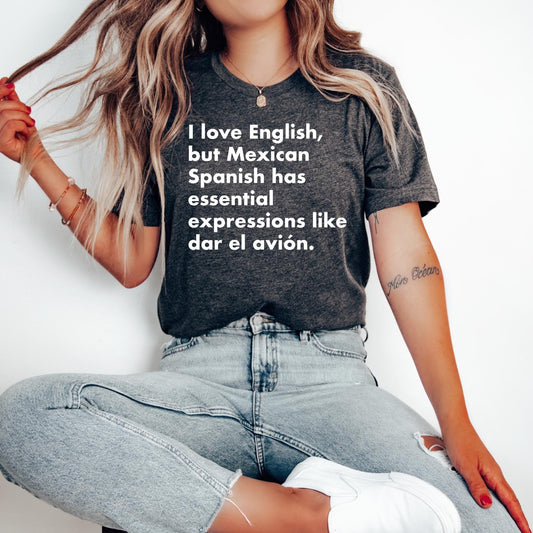 Latina woman sitting cross-legged wearing a heather gray t-shirt with the Spanish phrase "I love English, but Mexican Spanish has essential expressions like dar el avión."

